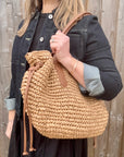 Woman wearing a black dress and denim jacket, holding a beige raffia shoulder handbag. 