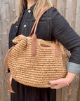 Woman wearing a black dress and denim jacket, holding a beige raffia shoulder handbag. 