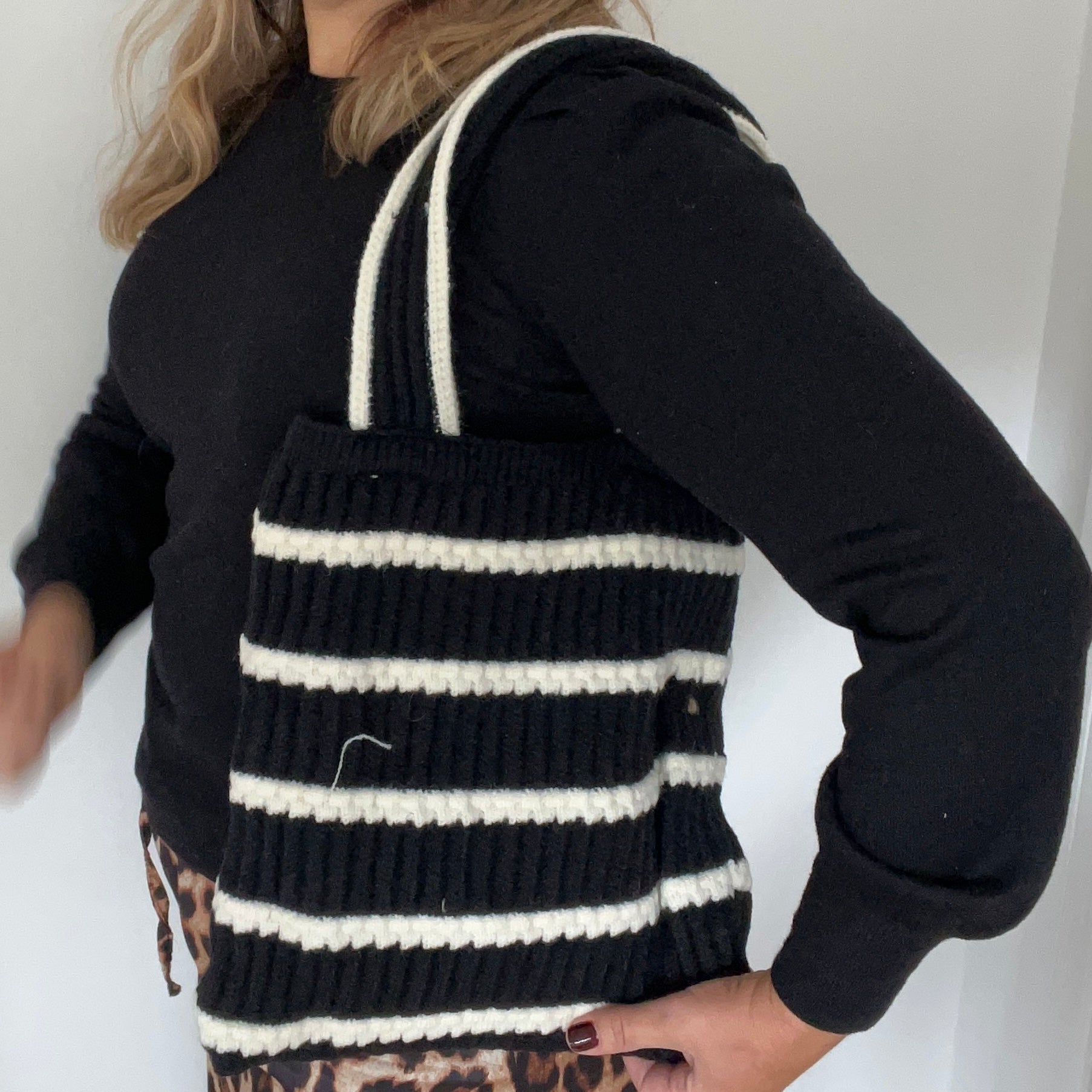 A knitted tote bag with black and cream stripes, featuring a double handle design, displayed on a person wearing a black sweater.