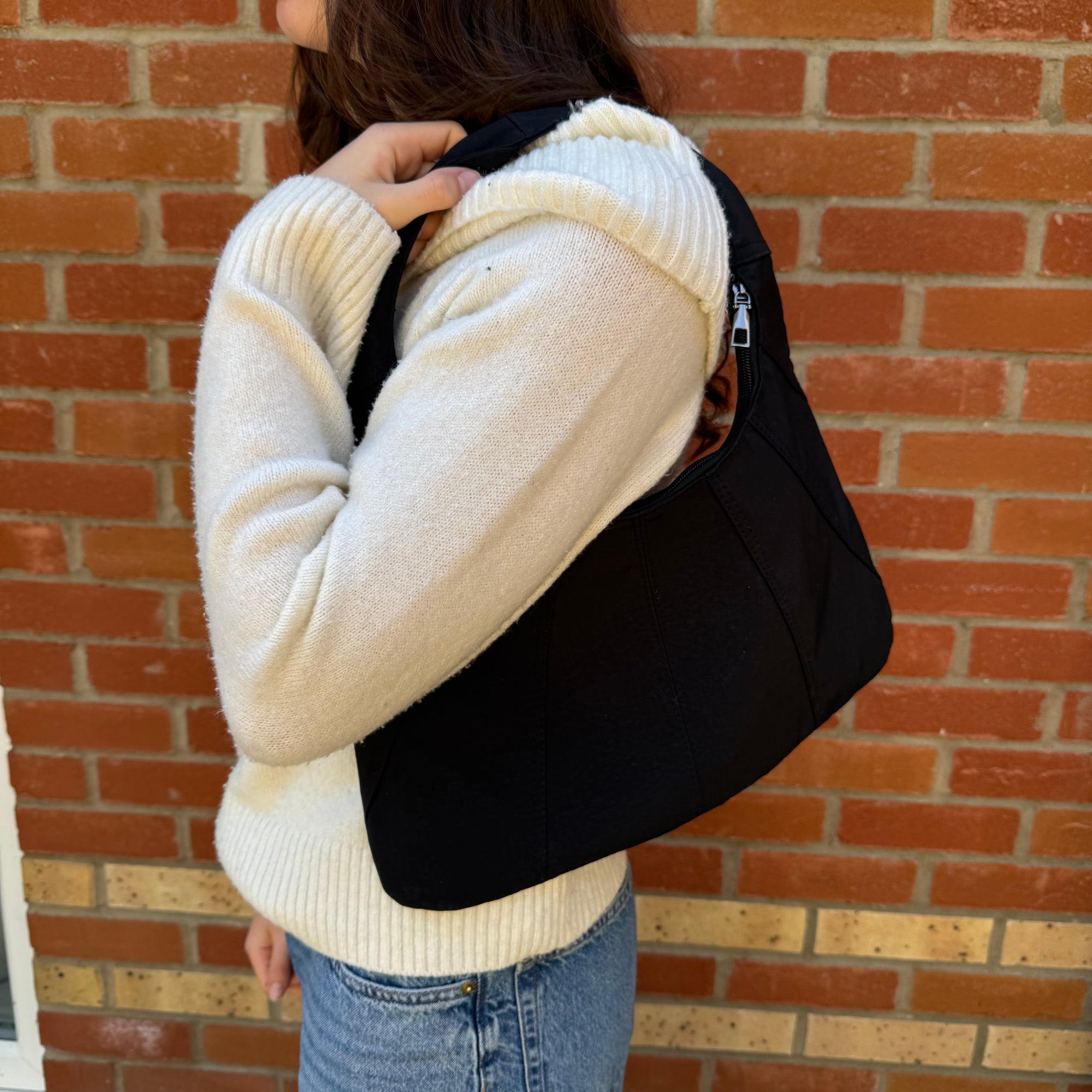 A woman wearing a beige sweater and blue jeans is holding a black fabric shoulder bag over their shoulder, standing against a brick wall background.