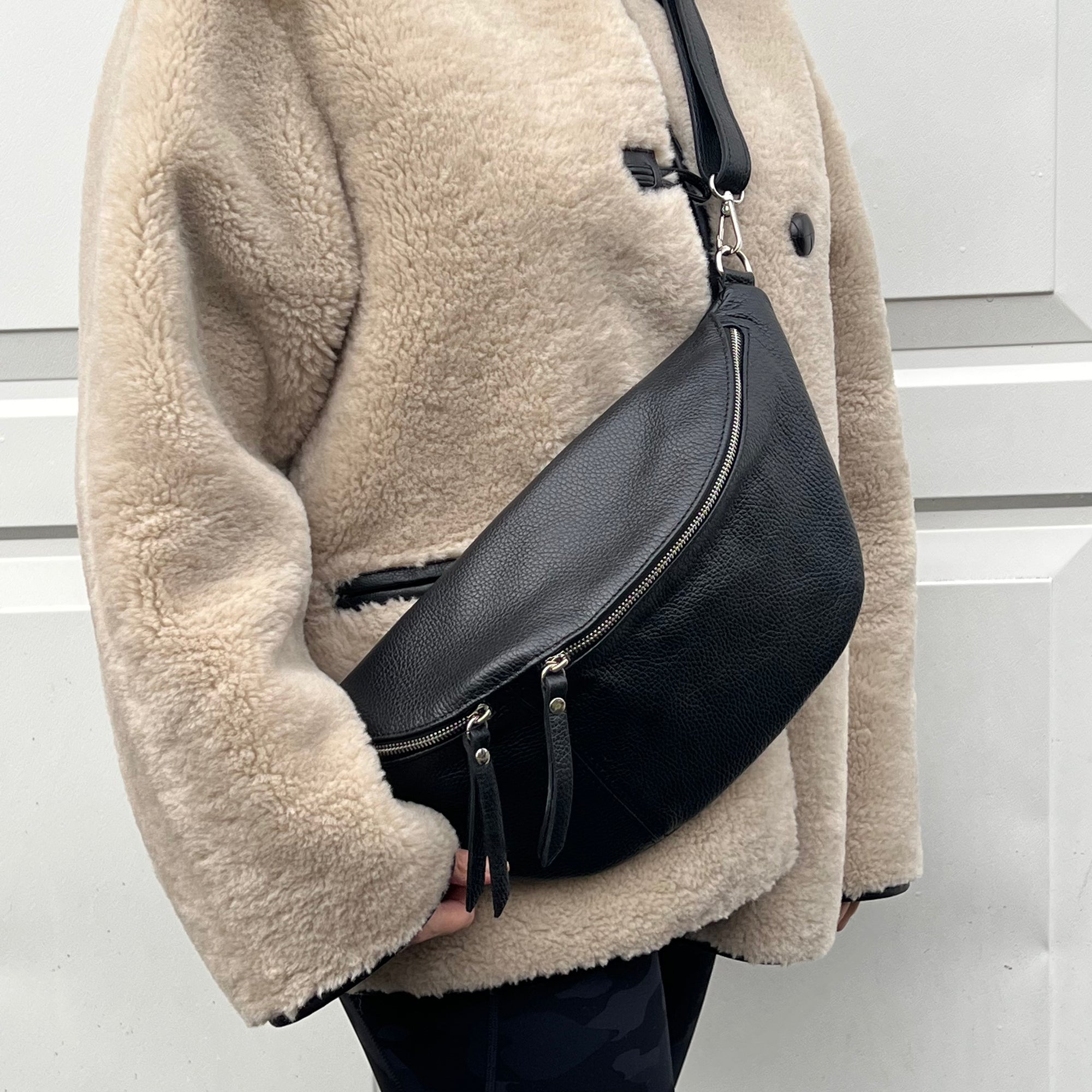 A person wearing a black large crossbody bum bag with an adjustable, detachable leather strap and multiple zip pockets.
