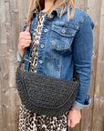 Woman wearing a denim jacket and leopard print top, holding a black raffia crossbody handbag. 