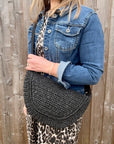 Woman wearing a denim jacket and leopard print top, holding a black raffia crossbody handbag. 
