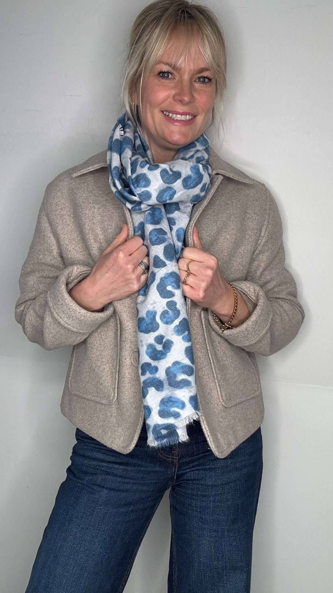 Woman wearing a beige jacket and blue jeans with a patterned scarf against a plain background