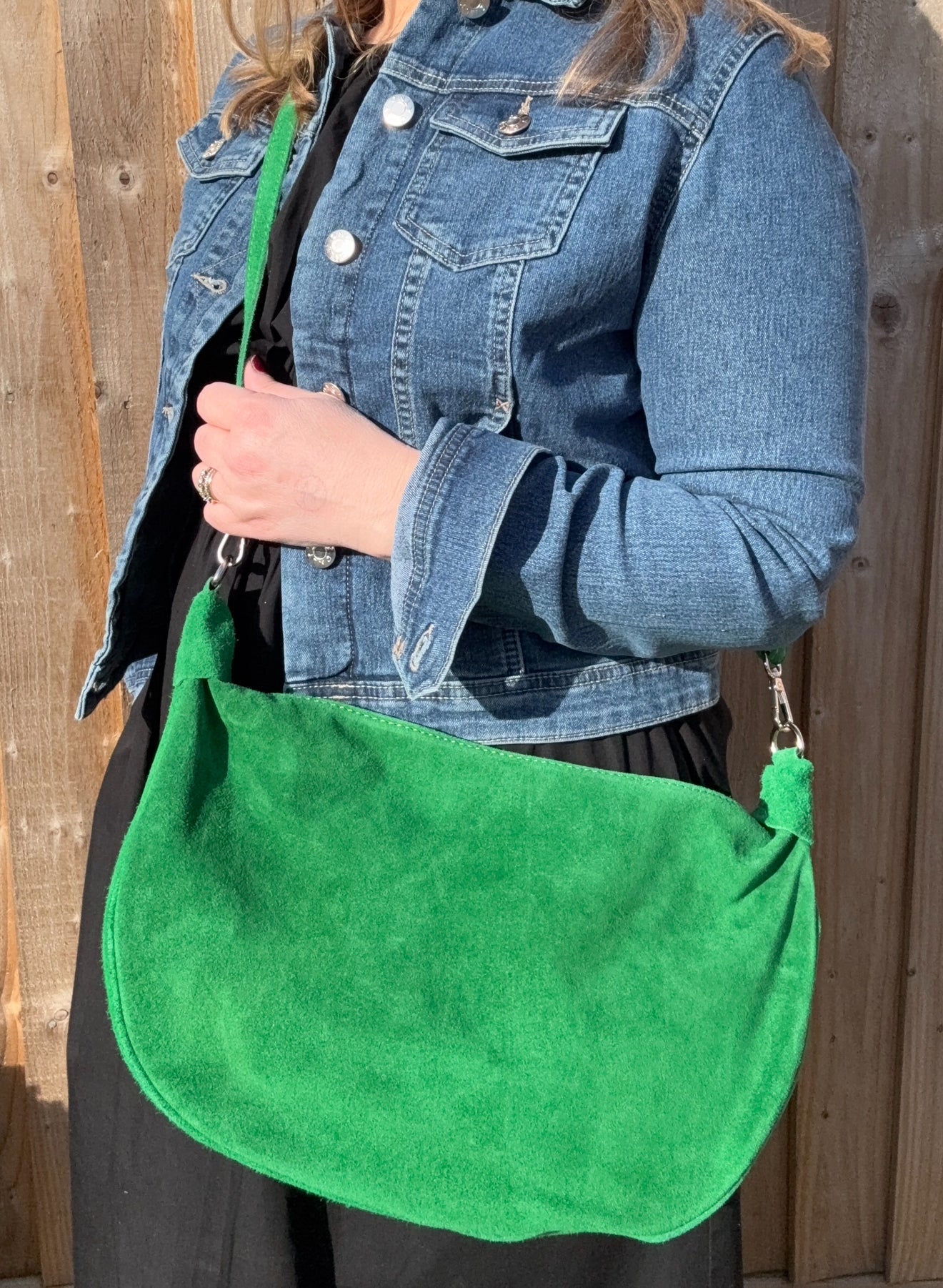 A woman wearing a denim jacket is holding a bright green large suede crossbody swing bag with a green-colored chain strap.
