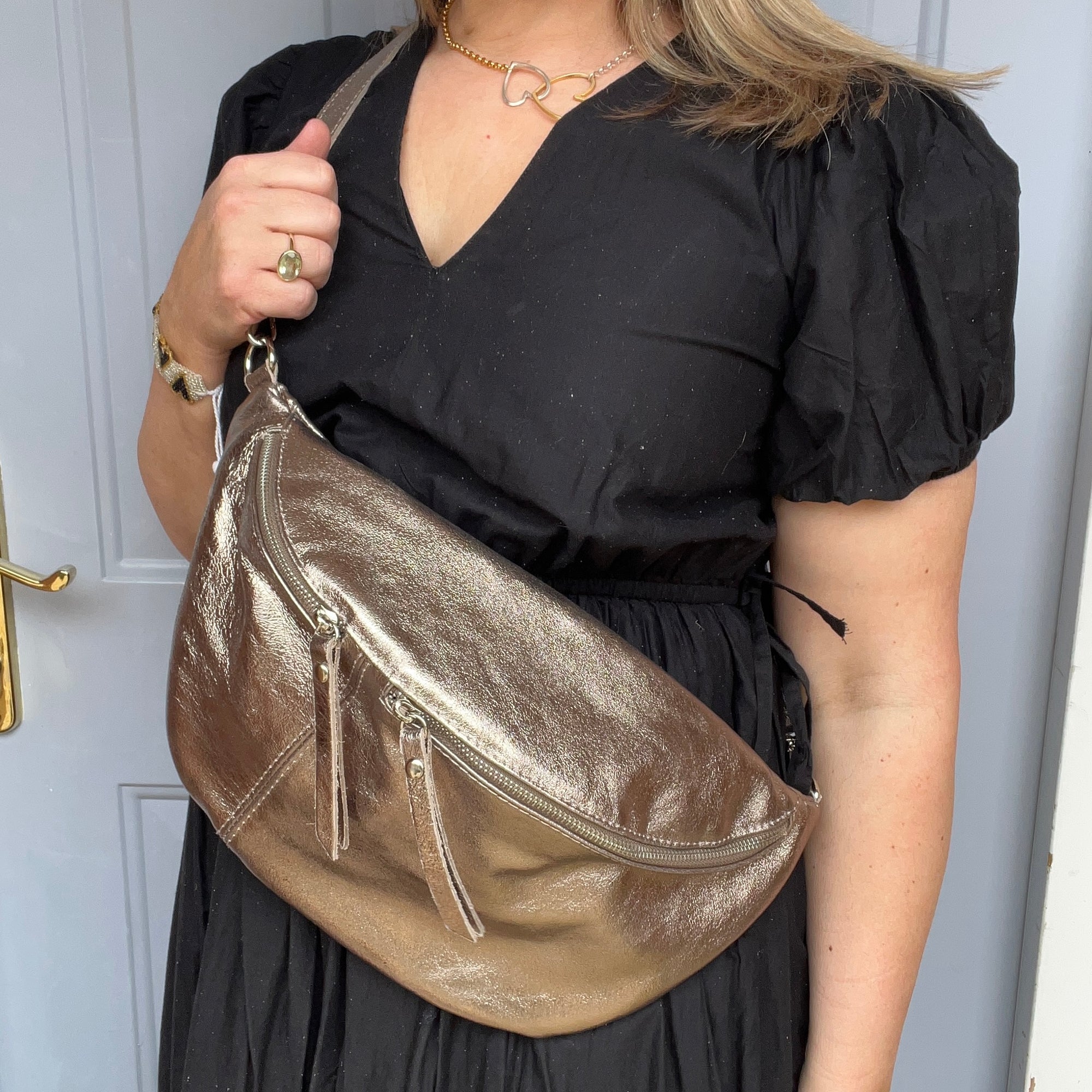 
a woman wearing a black dress holding a large bronze crossbody bum bag. 
