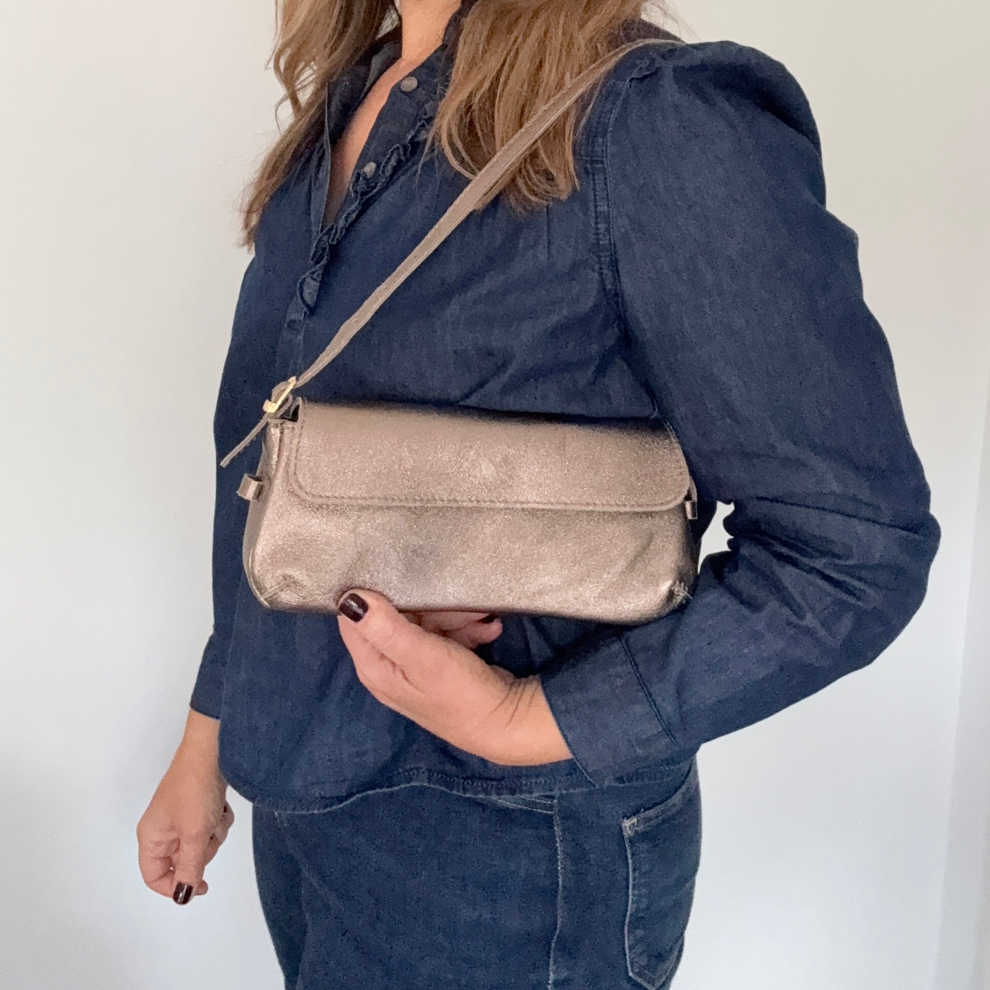 Woman wearing a denim jacket holding a bronze clutch handbag against a white background.