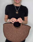 Woman wearing a black dress, holding a brown woven crossbody handbag.