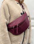 A woman wearing a beige coat and a burgundy large crossbody bum bag with a gold zipper.