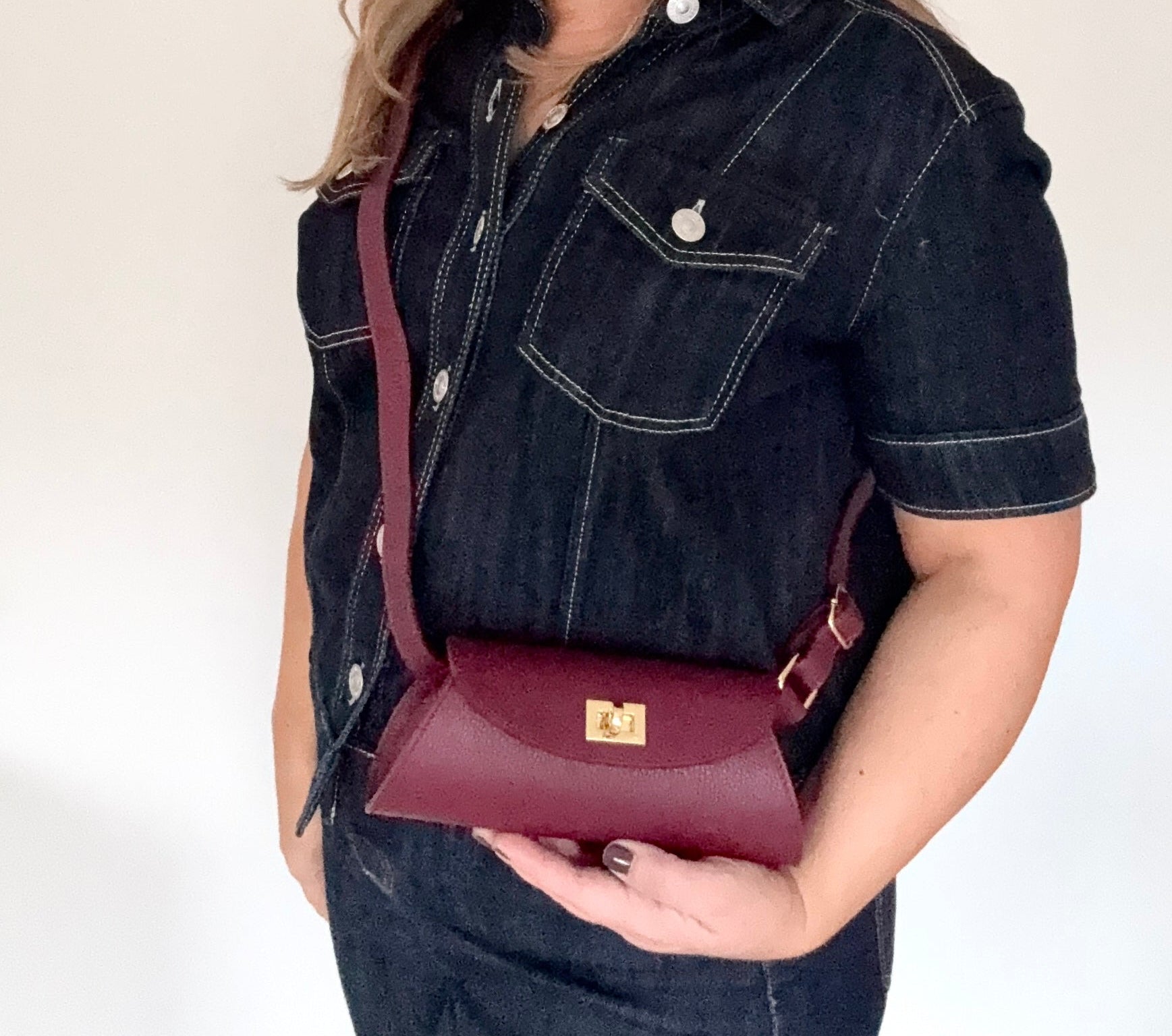 Woman wearing a dark denim shirt holding a burgundy leather buckle detail crossbody handbag 