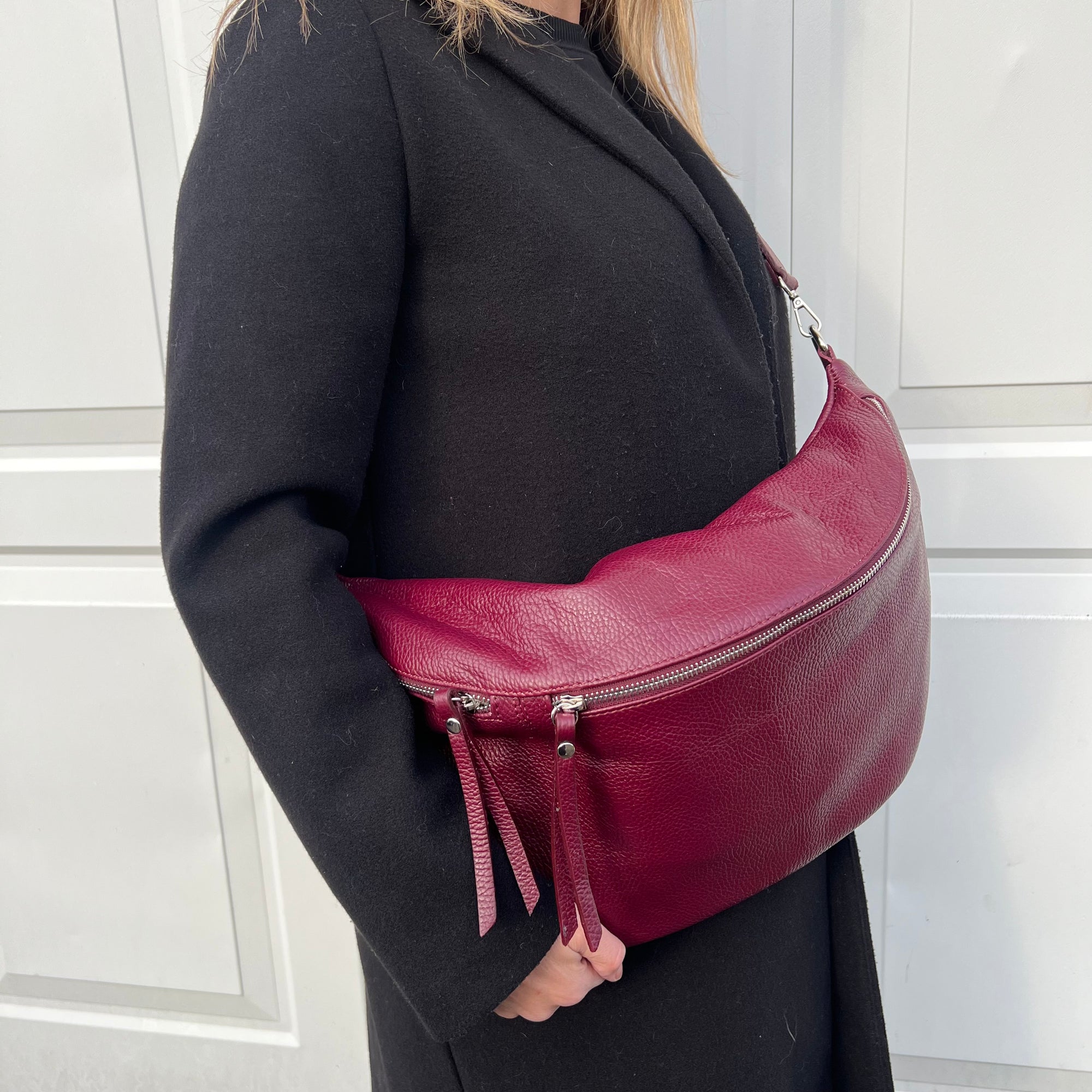 A person wearing a black coat is carrying a burgundy XL leather crossbody bag with silver hardware.
