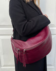 A person wearing a black coat is carrying a burgundy XL leather crossbody bag with silver hardware.  