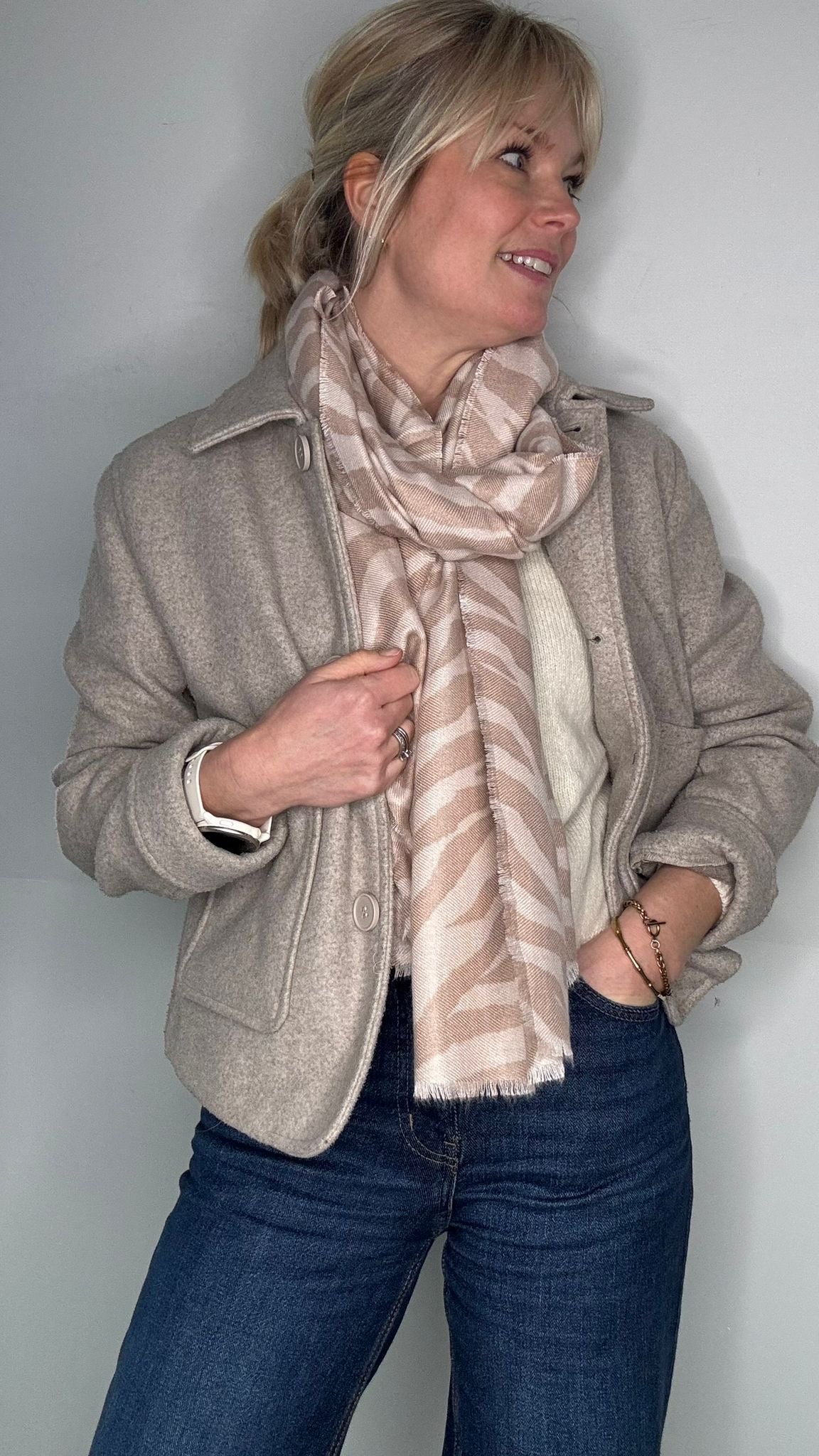 Woman wearing a beige jacket and a camel and beige scarf, and blue jeans against a plain background