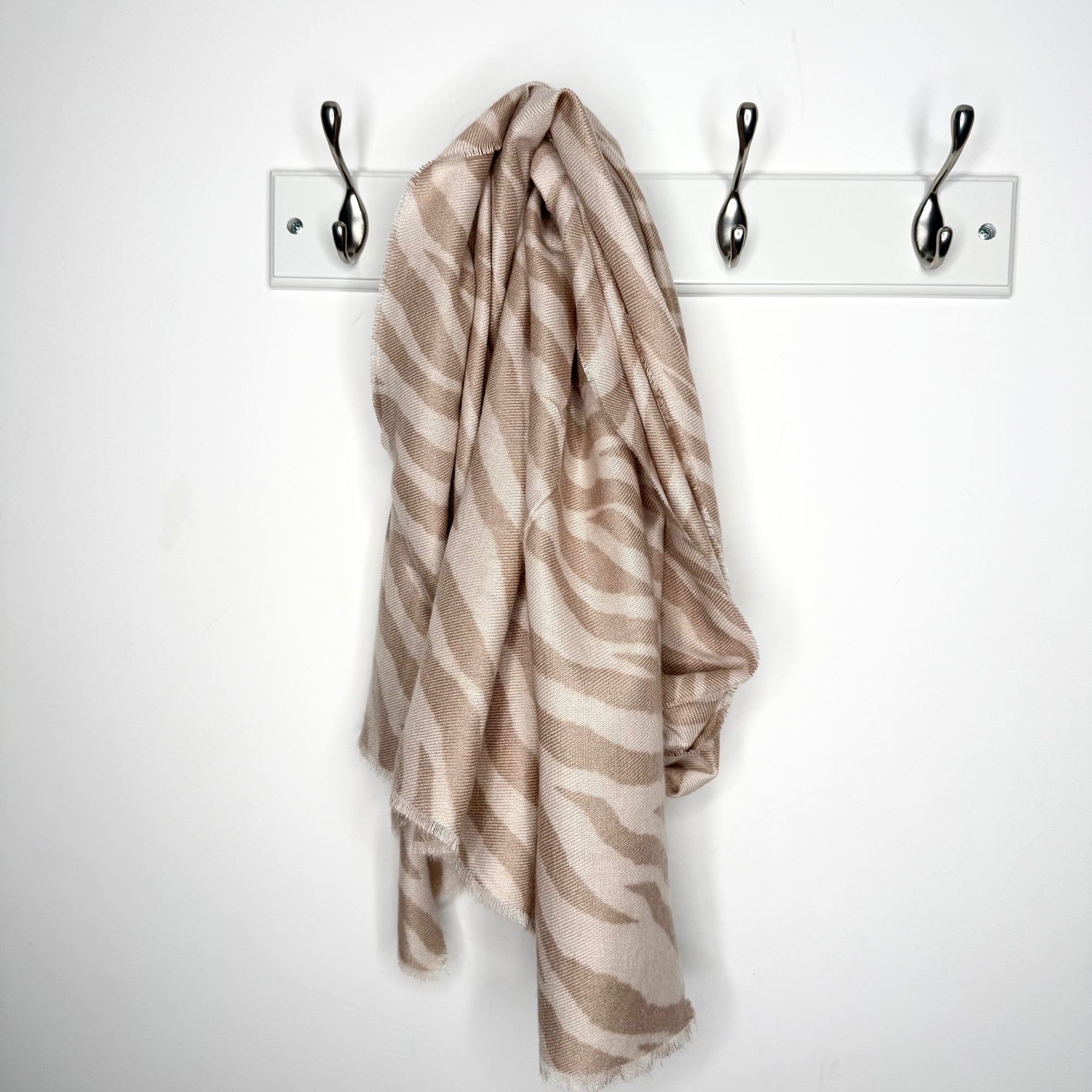 Beige and camel patterned scarf hanging on a white rack with three hooks against a white background.