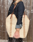 Woman wearing a denim jacket, holding a large cream and beige raffia tote bag. 