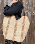 Woman wearing a denim jacket, holding a large cream and beige raffia tote bag. 