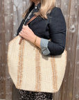 Woman wearing a denim jacket, holding a large cream and beige raffia tote bag. 