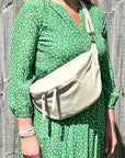 A person wearing a cream-colored large crossbody bum bag with an adjustable, detachable leather strap and multiple zip pockets, carried over the shoulder.
