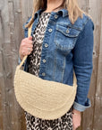 Woman wearing a denim jacket and leopard print top, holding a cream raffia crossbody handbag. 