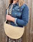 Woman wearing a denim jacket and leopard print top, holding a cream raffia crossbody handbag. 