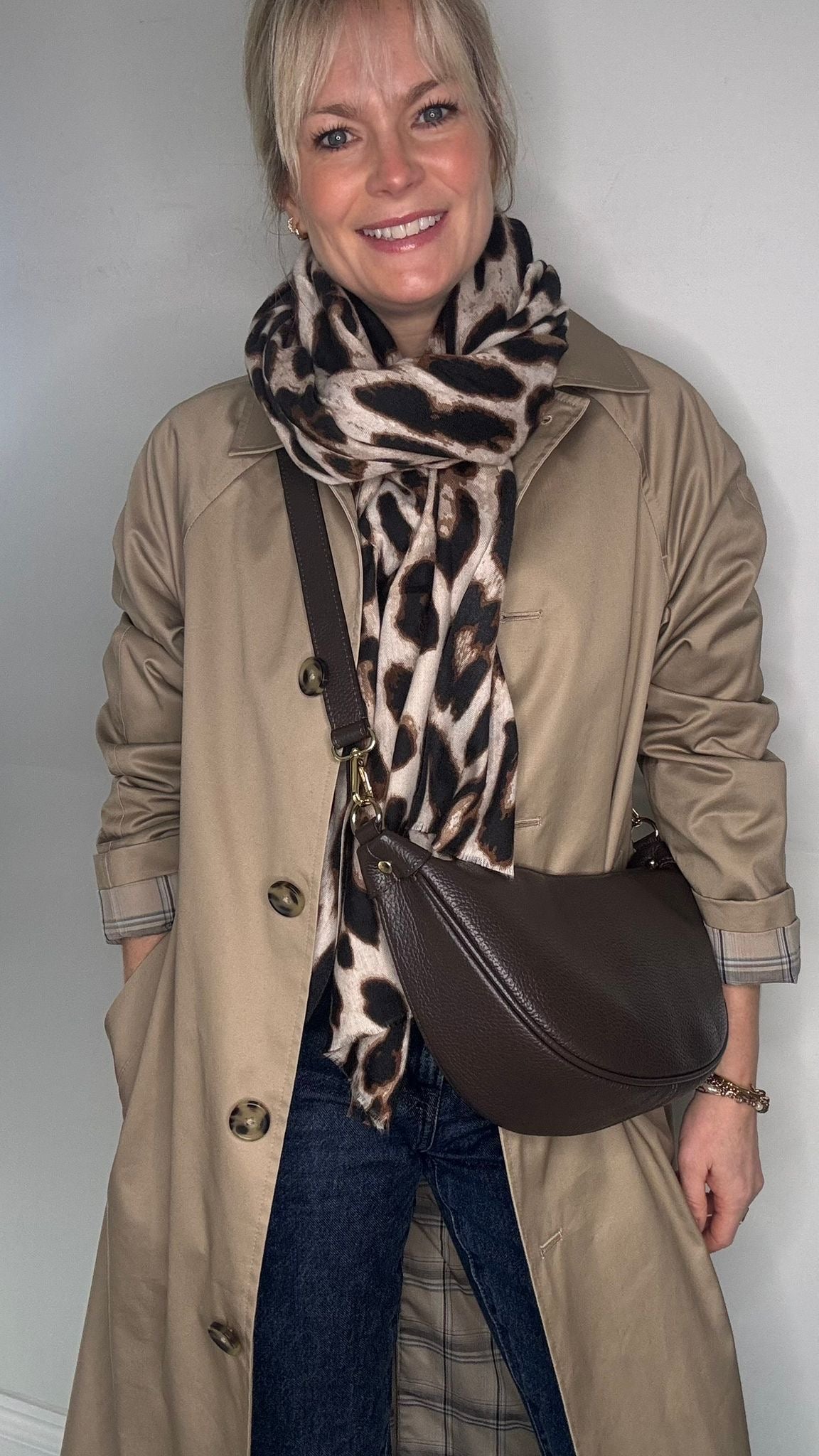 Person wearing a beige trench coat, blue jeans, and a brown crossbody  bag with a leopard print scarf.
