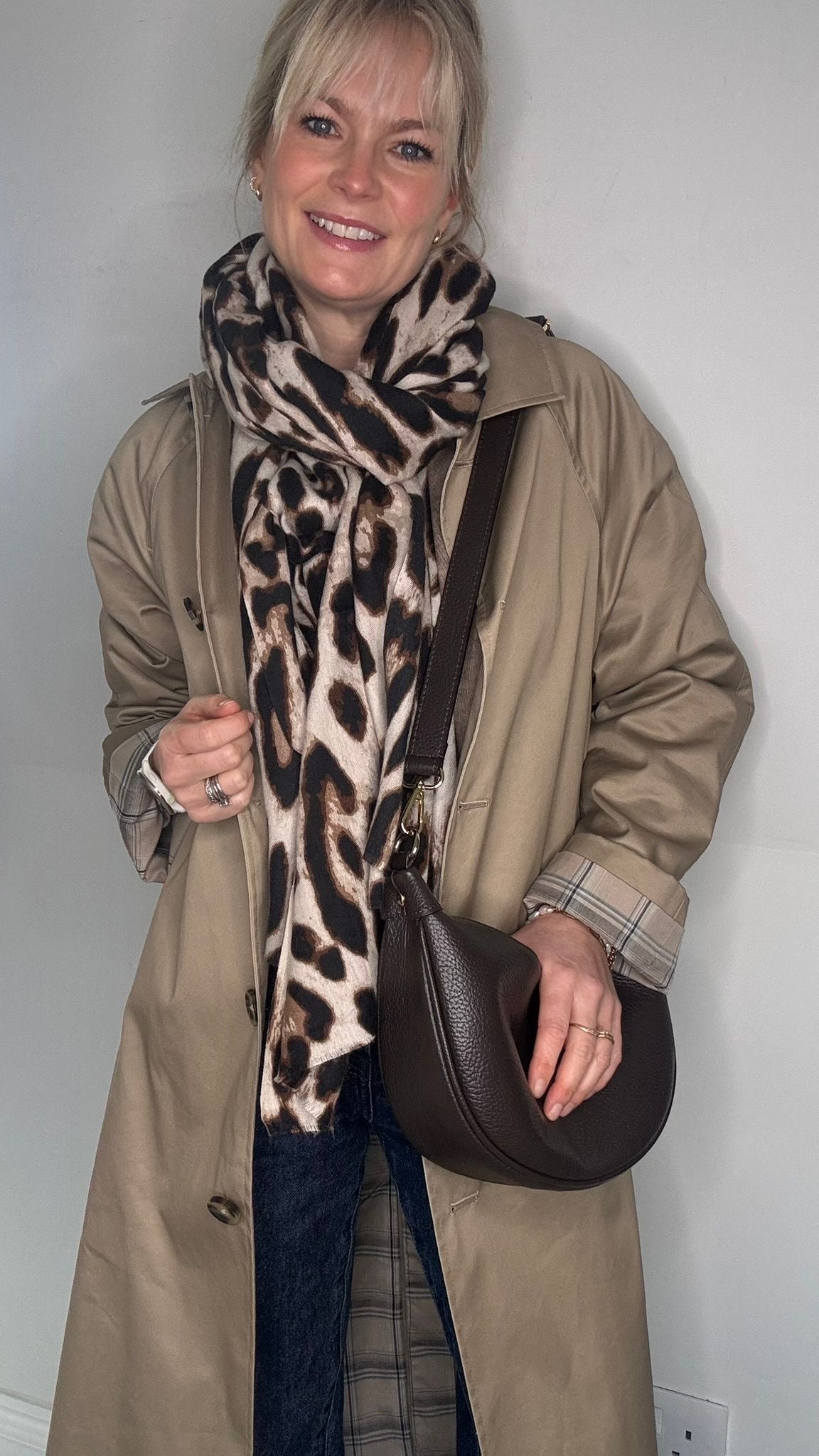 Woman wearing a beige trench coat, leopard print scarf, and holding a brown crossbody bag 