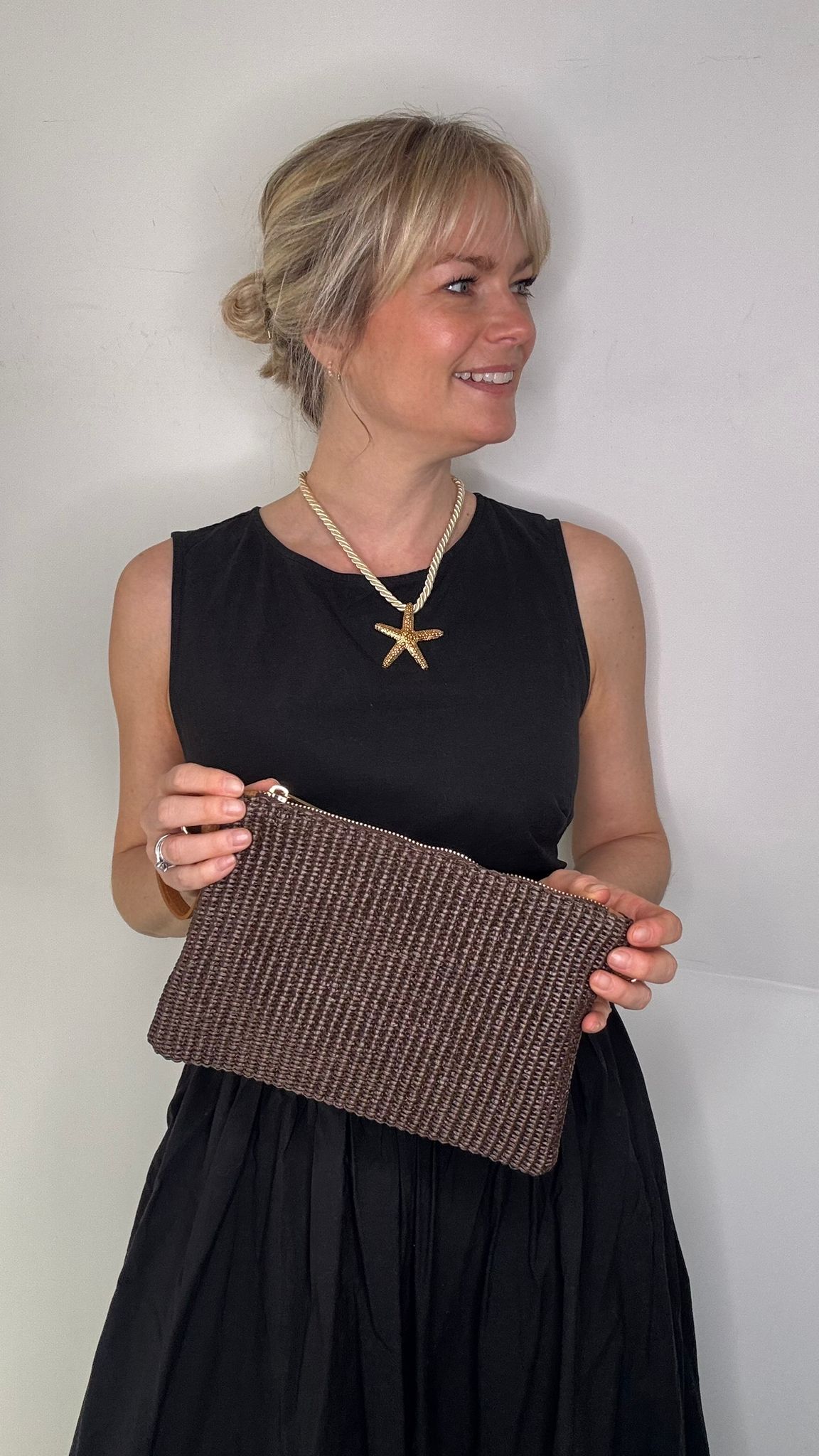 Woman wearing a black dress, holding a dark brown clutch / crossbody handbag. 