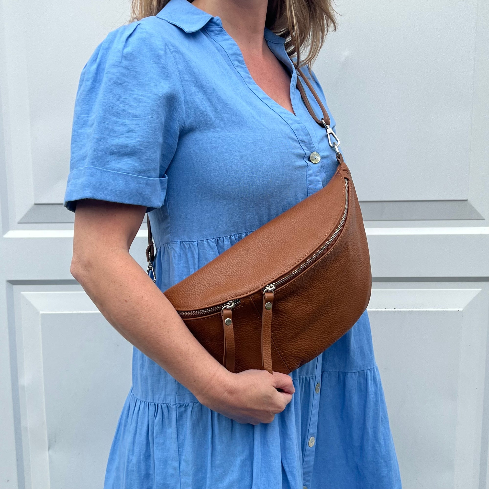 A person wearing a blue dress is holding a dark tan large crossbody bum bag with an adjustable strap and silver hardware.