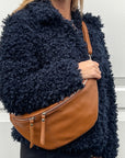 A woman  wearing a dark tan large crossbody bum bag with an adjustable, detachable leather strap with silver hardware. 