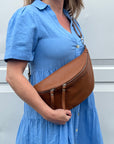 A person wearing a blue dress is holding a dark tan large crossbody bum bag with an adjustable strap and silver hardware.