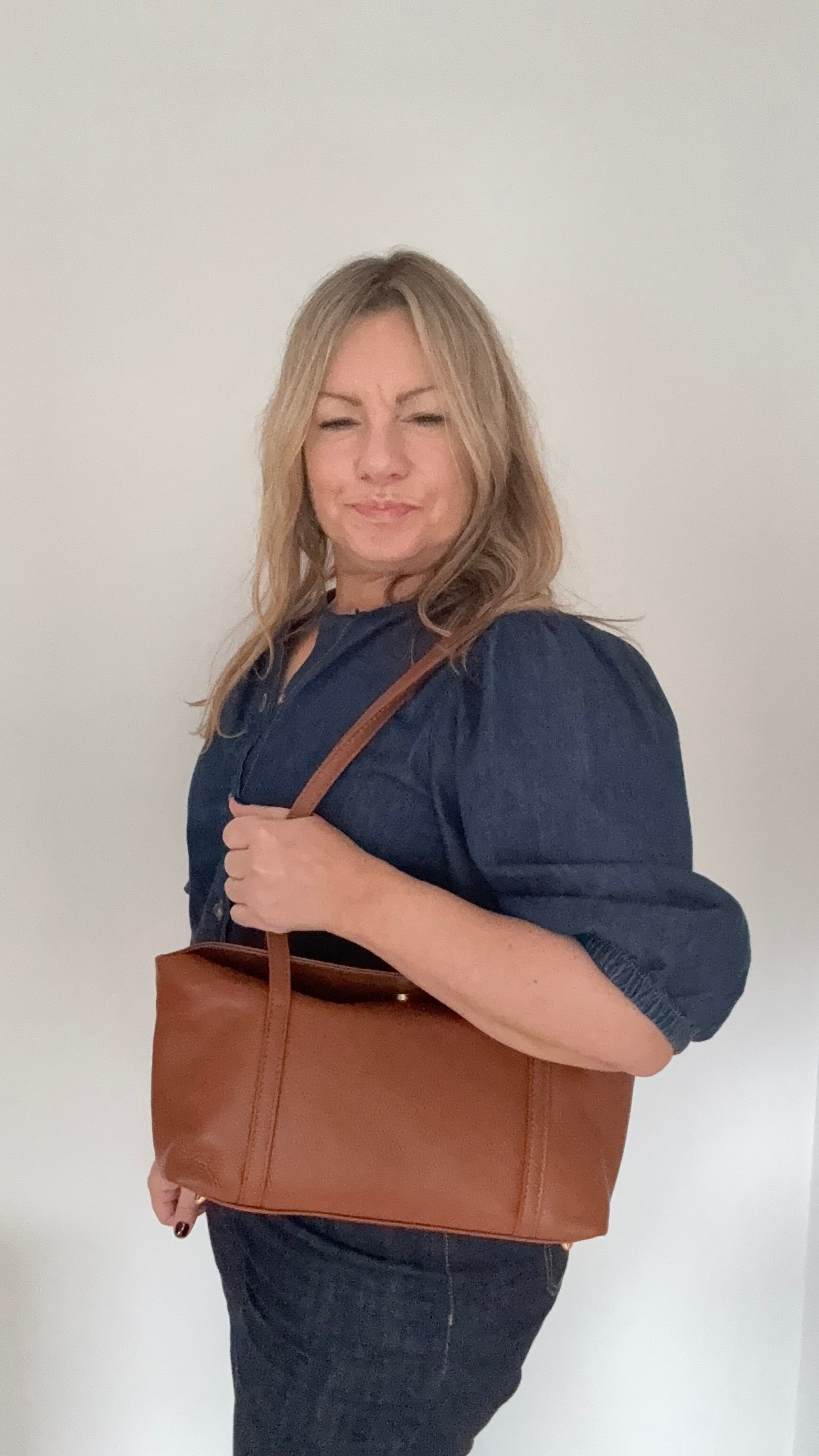 woman with blond hair and a denim shirt is  holding a dark tan leather  shoulder handbag 