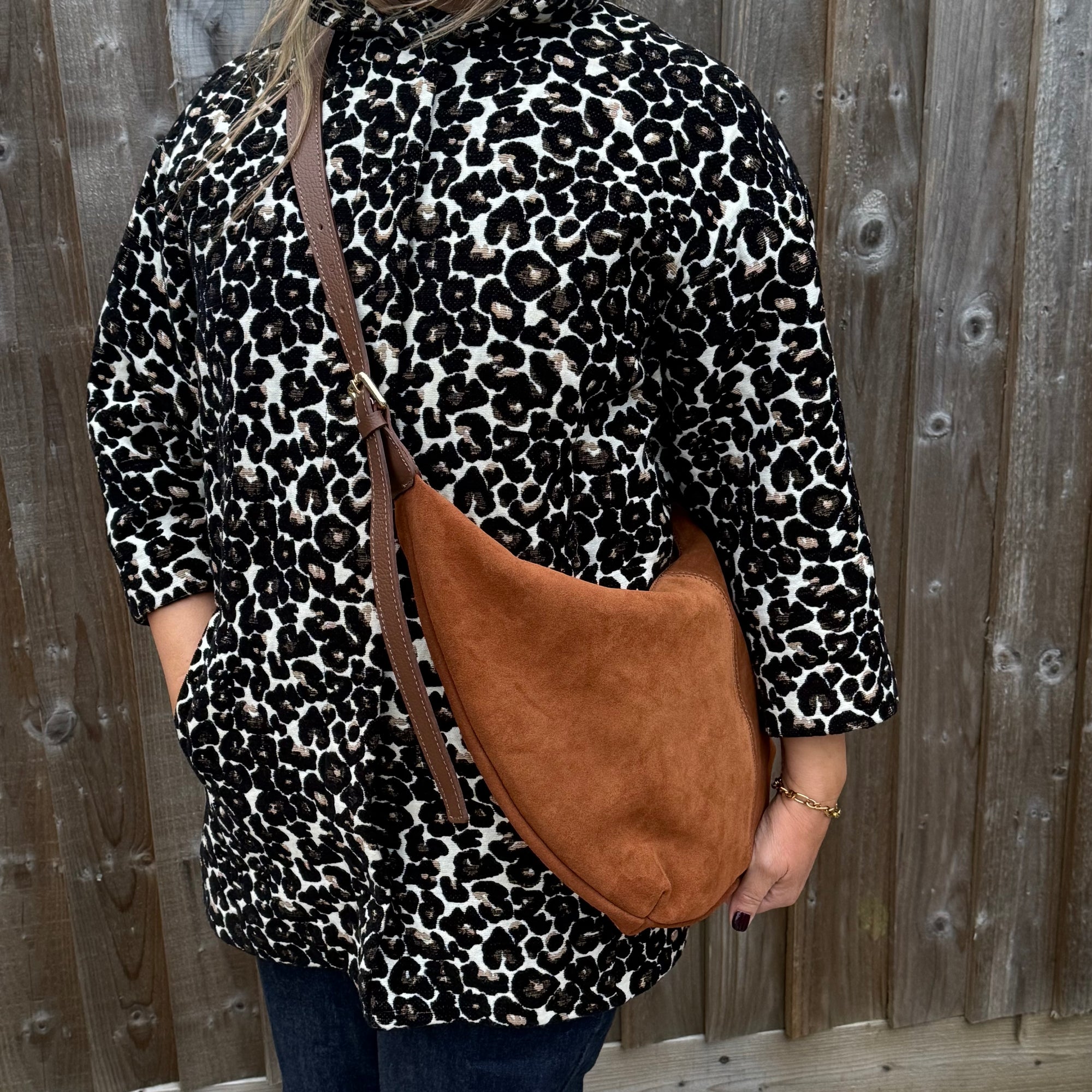woman wearing a leopard print shirt with a dark tan XL crossbody bag against a wooden fence background
