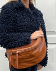 A woman  wearing a dark tan XL leather crossbody bum bag with two front zip openings and an adjustable strap.