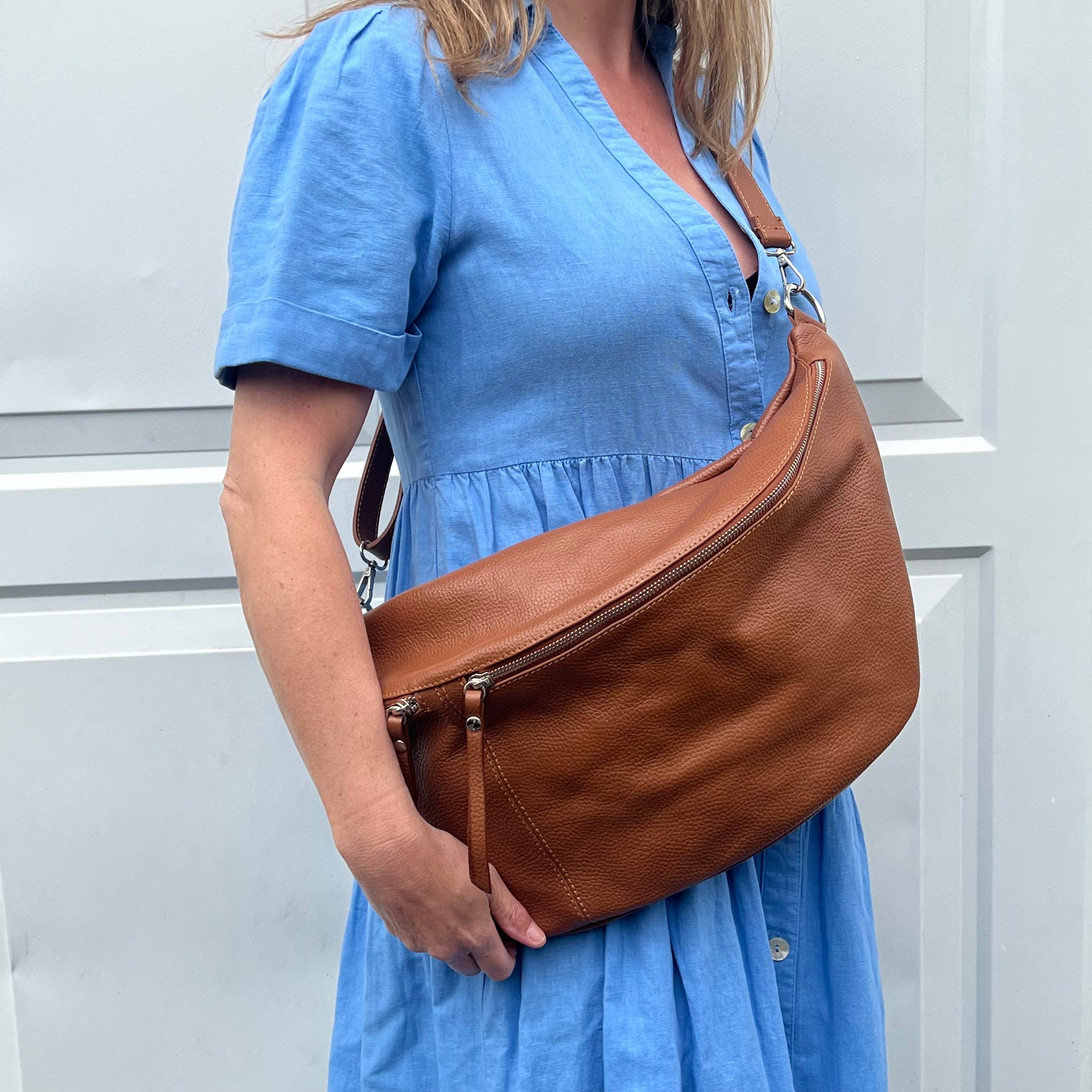 A woman  wearing a dark tan XL leather crossbody bum bag with two front zip openings and an adjustable strap.

