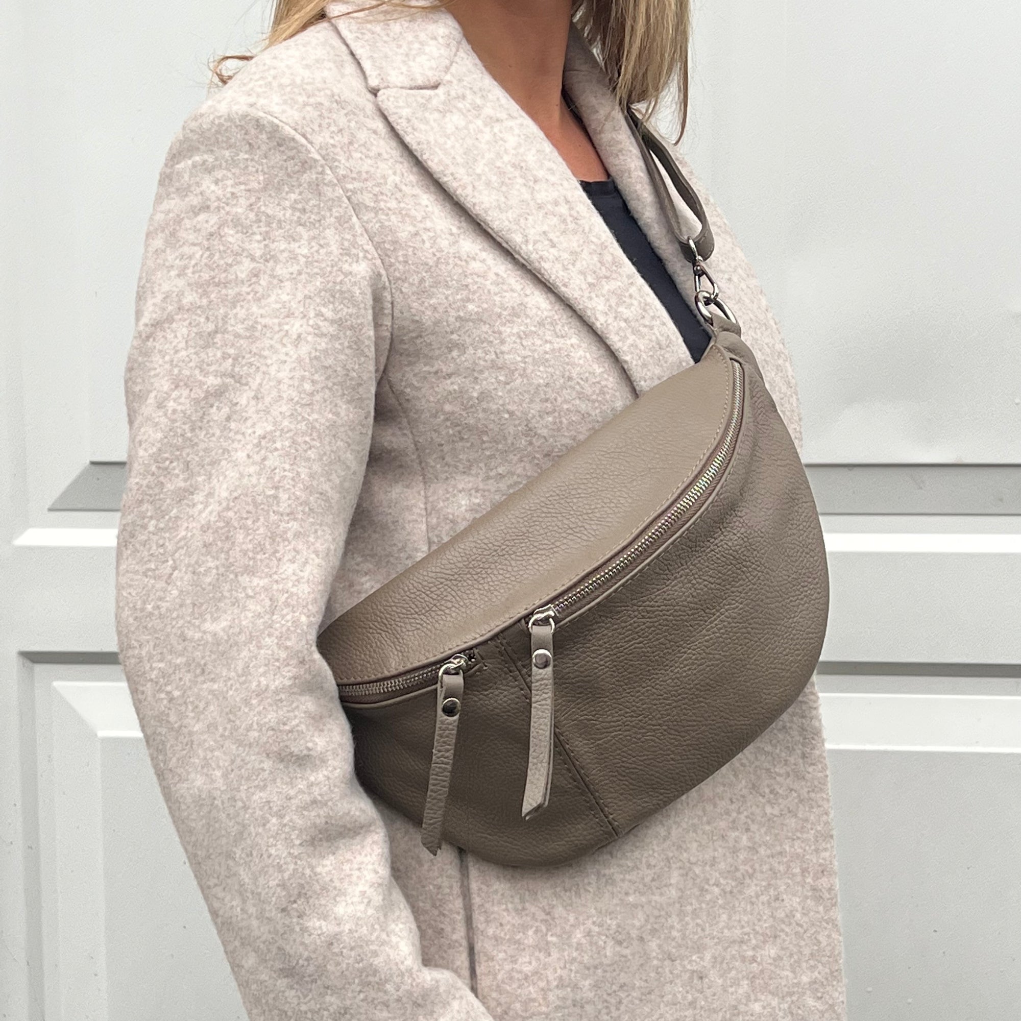 A woman wearing a dark taupe large crossbody bum bag with an adjustable, detachable leather strap and multiple zip pockets.