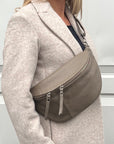 A woman wearing a dark taupe large crossbody bum bag with an adjustable, detachable leather strap and multiple zip pockets.