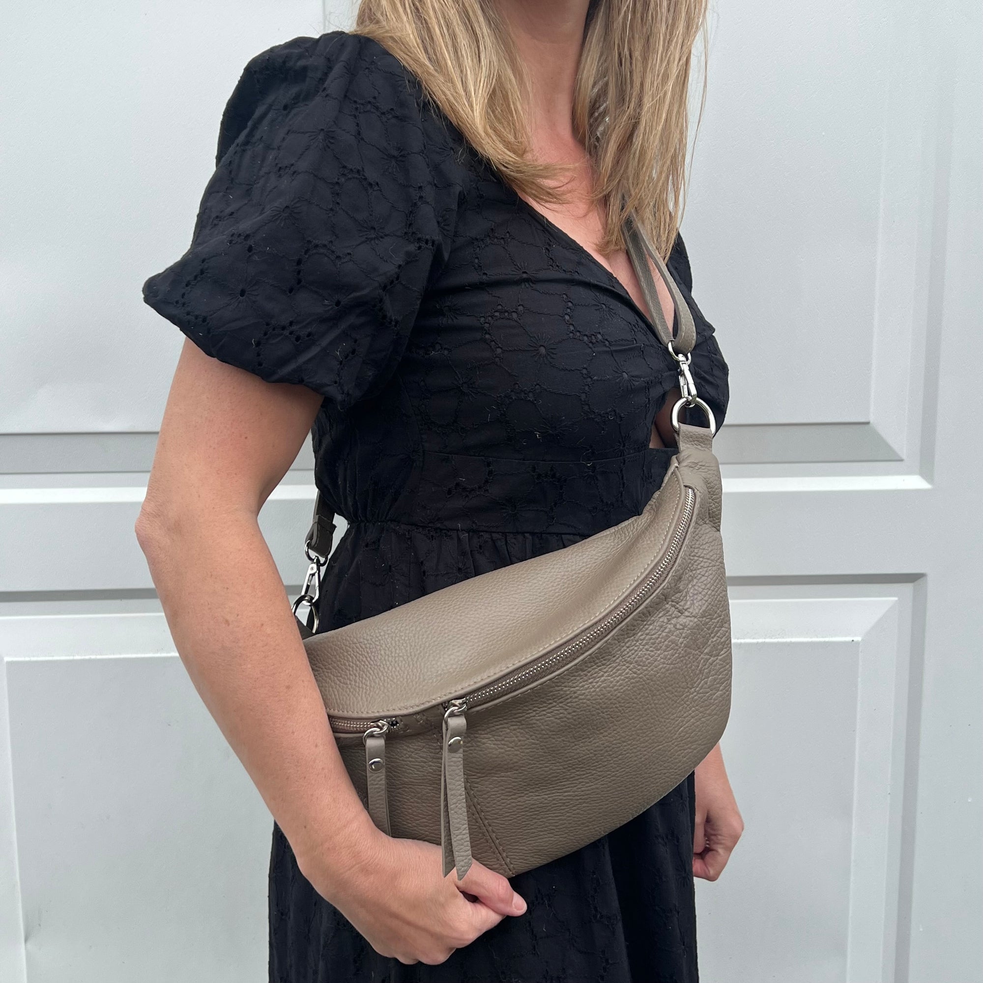 A person wearing a dark taupe large crossbody bum bag with an adjustable leather strap and multiple zip pockets.