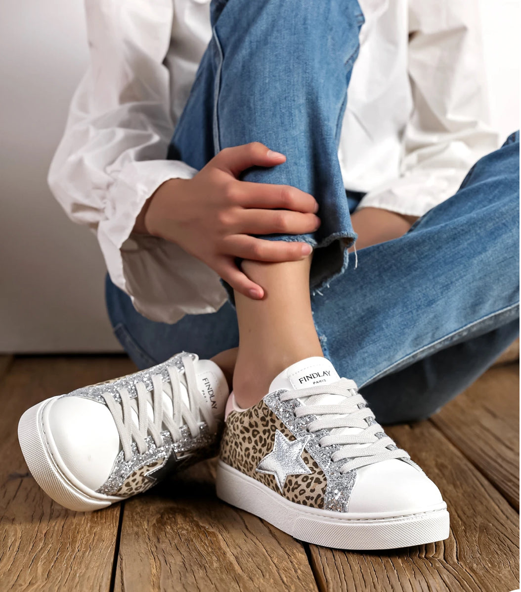 A person seated on wooden flooring wearing blue jeans and white leopard print & star lace-up trainers with a silver star and pale pink heel detail.