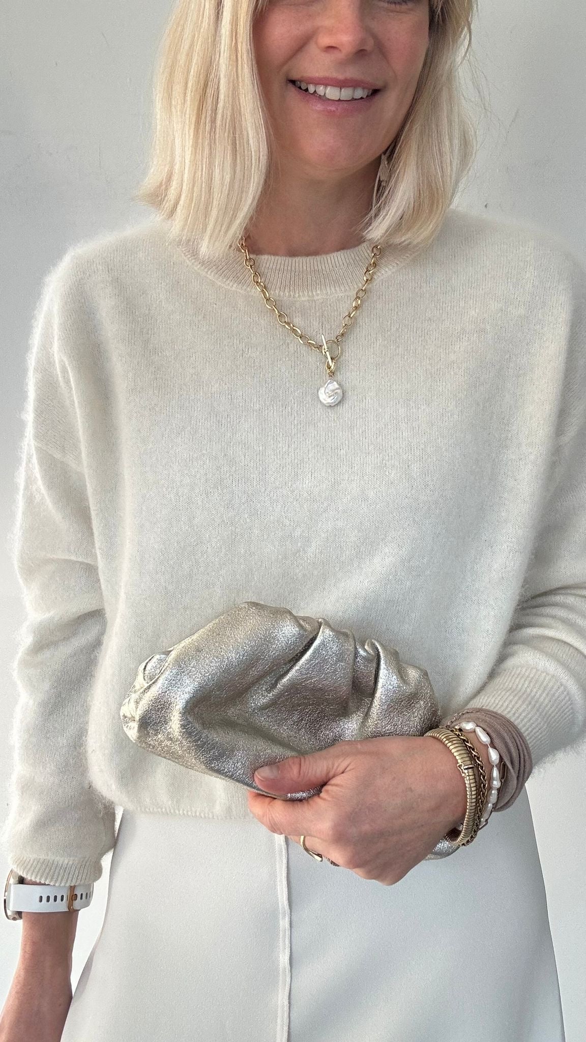 A woman  wearing a light grey sweater holding a silver clutch against a neutral background
