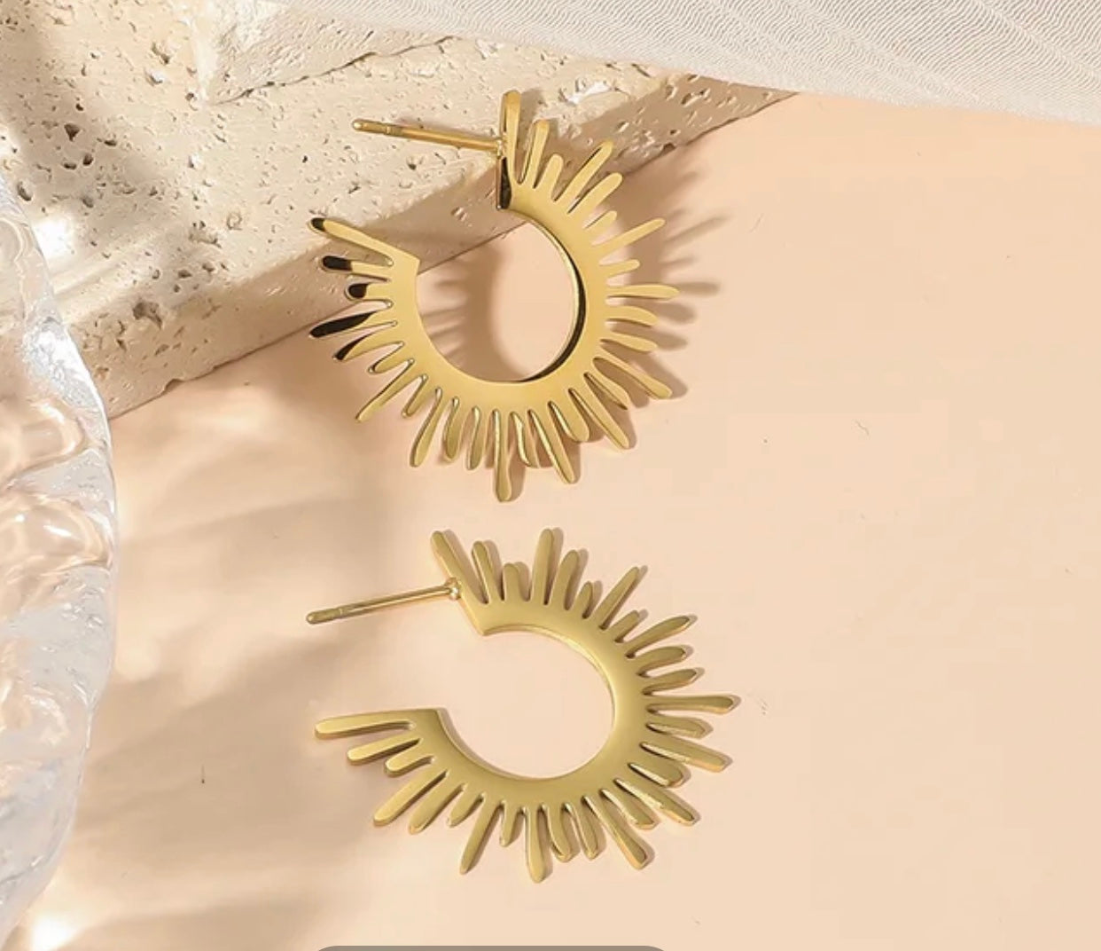 Gold hoop earrings with a sunburst design on a beige surface.