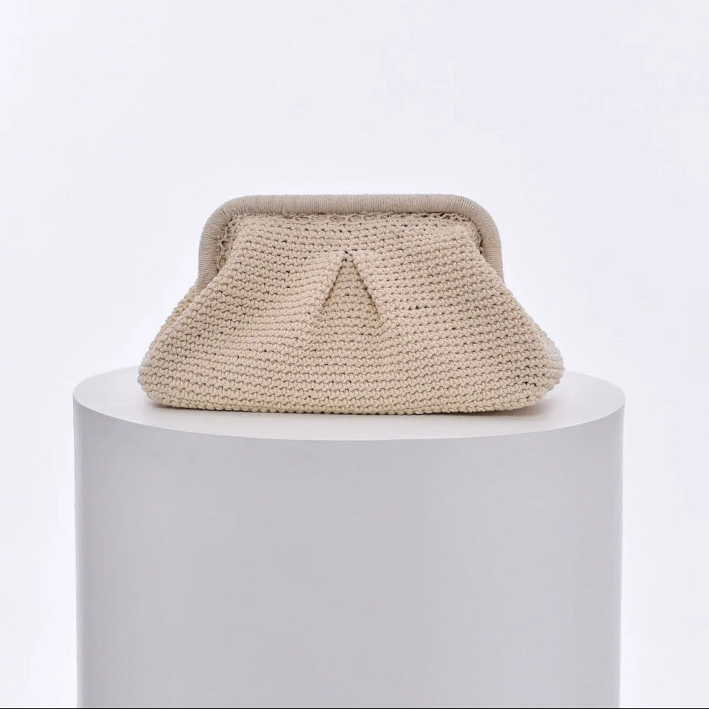 Cream knitted cloud clutch bag with a triangle design on a white pedestal against a light gray background