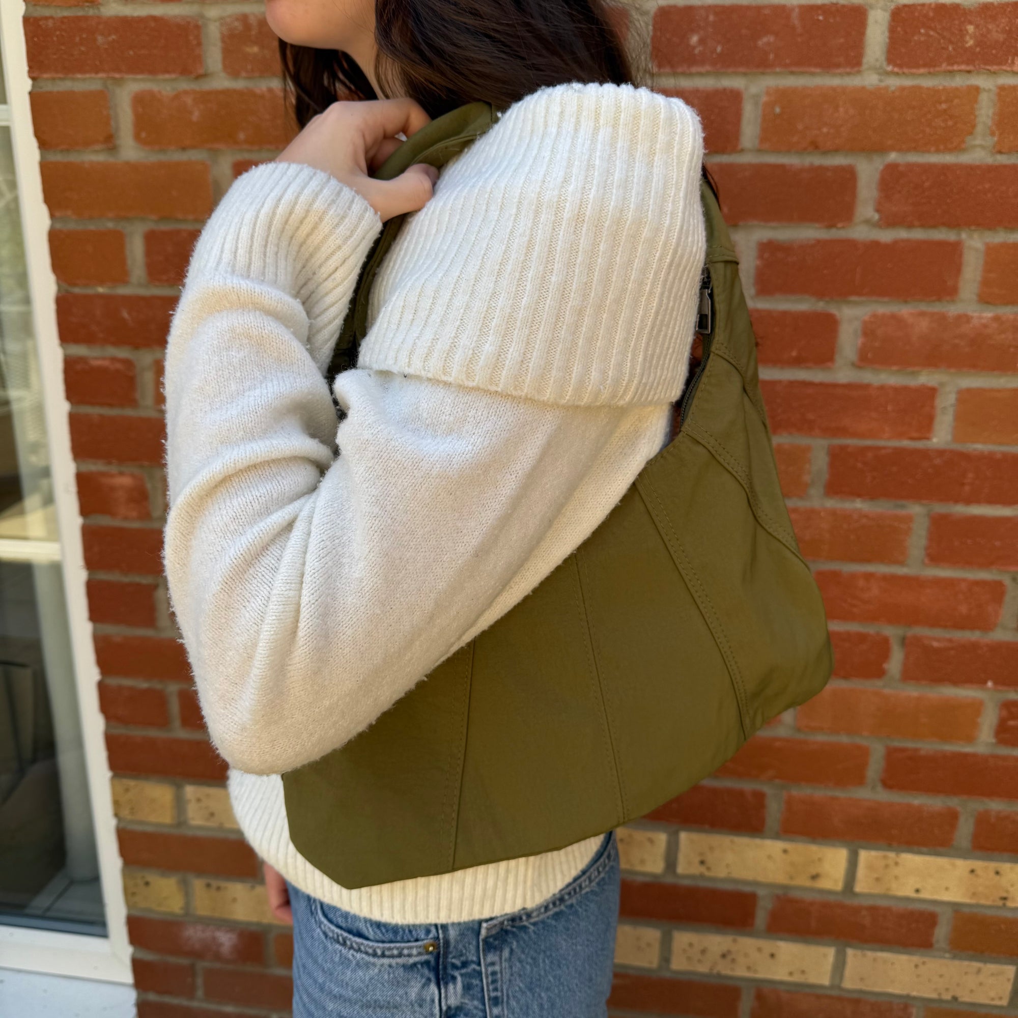 A woman wearing a beige sweater carrying a khaki fabric shoulder bag over their shoulder.