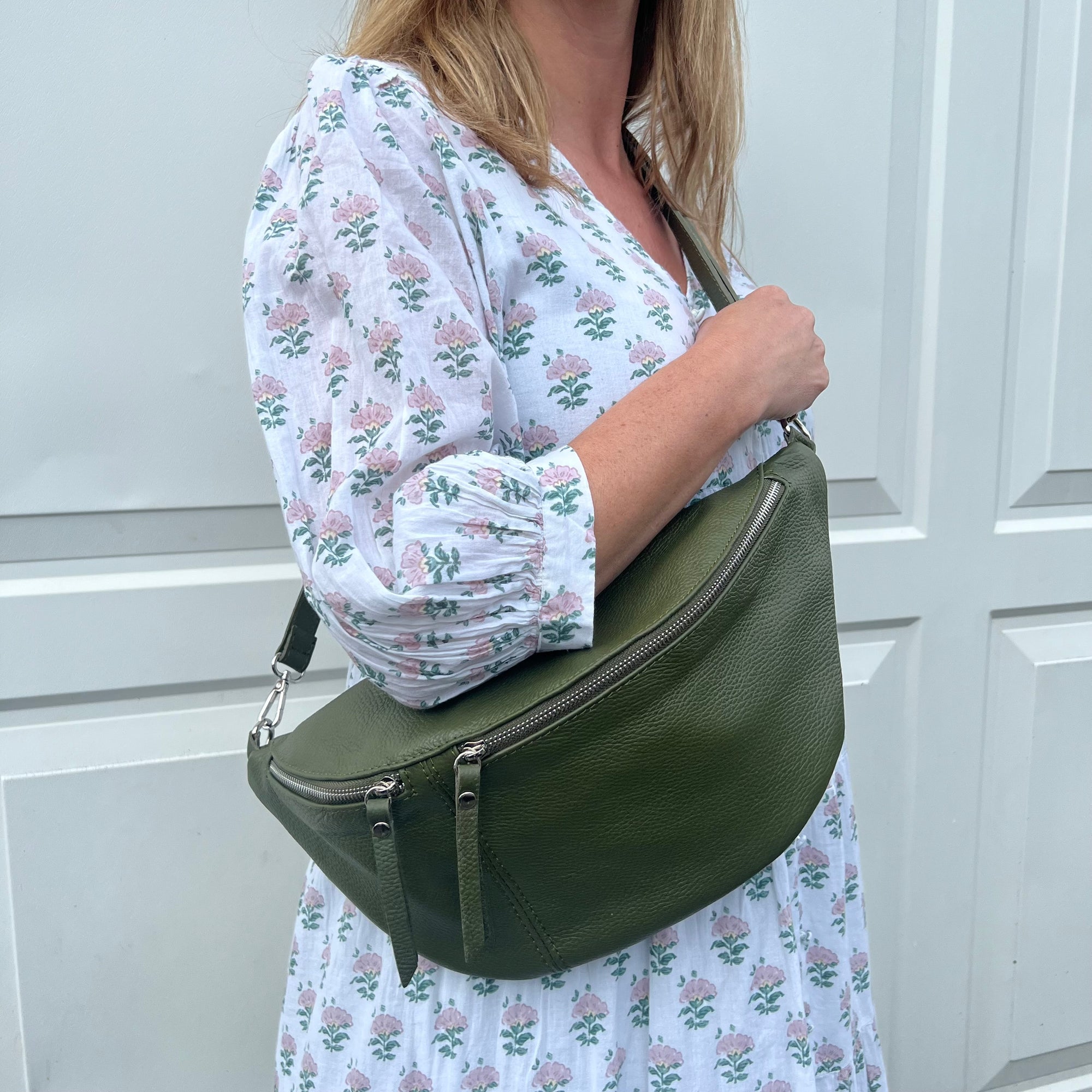 A woman wearing a khaki crossbody bum bag with silver accented hardware. 
