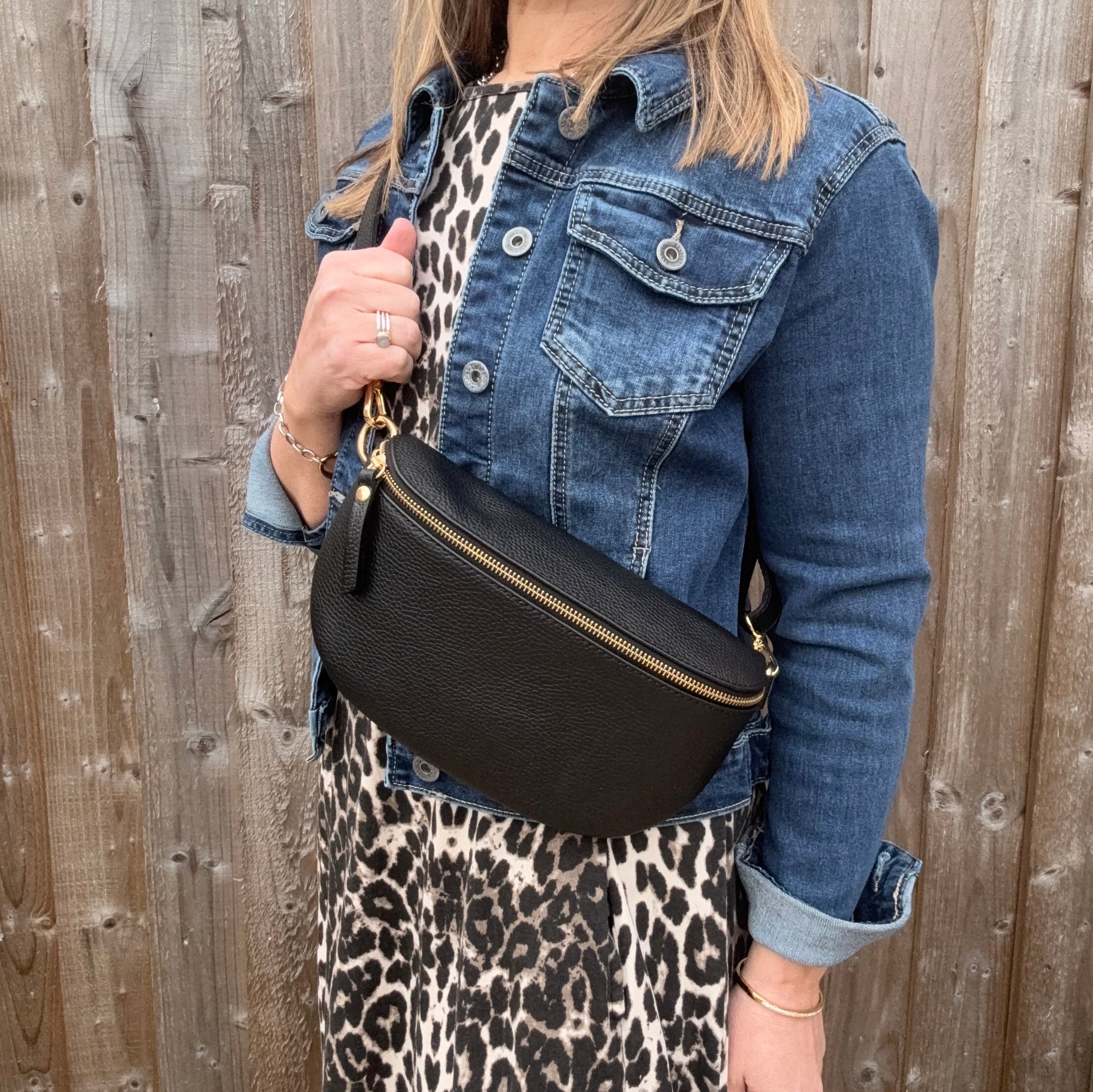 Woman wearing a denim jacket and leopard print skirt holding a large black crossbody/ waist bag against a wooden fence.