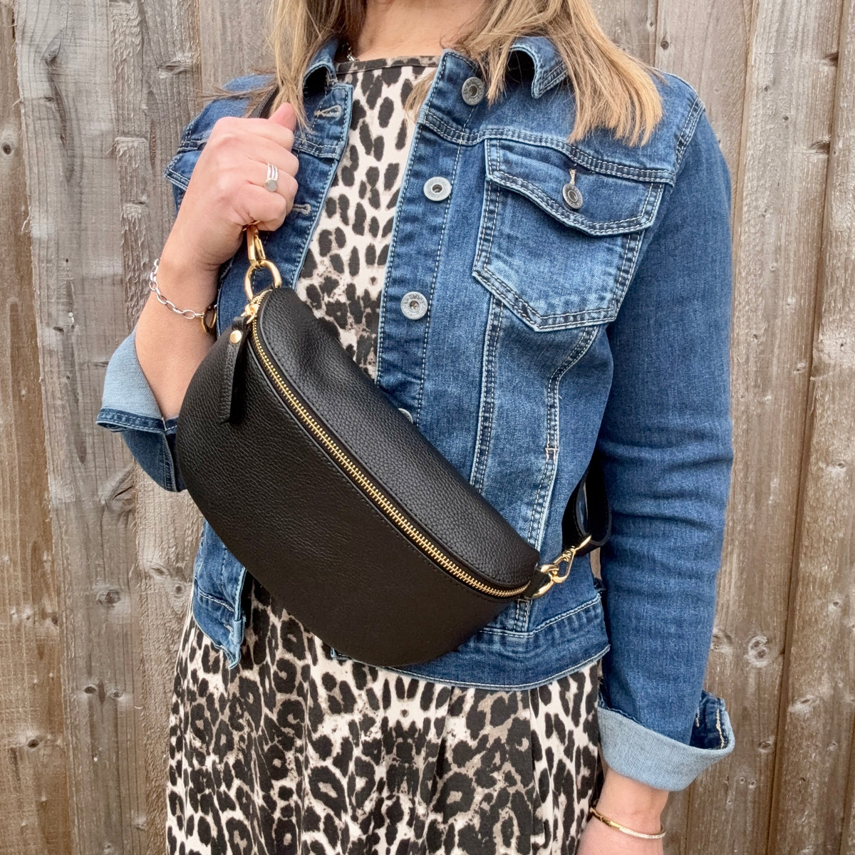 Woman wearing a denim jacket and leopard print skirt holding a large black crossbody/ waist bag against a wooden fence.