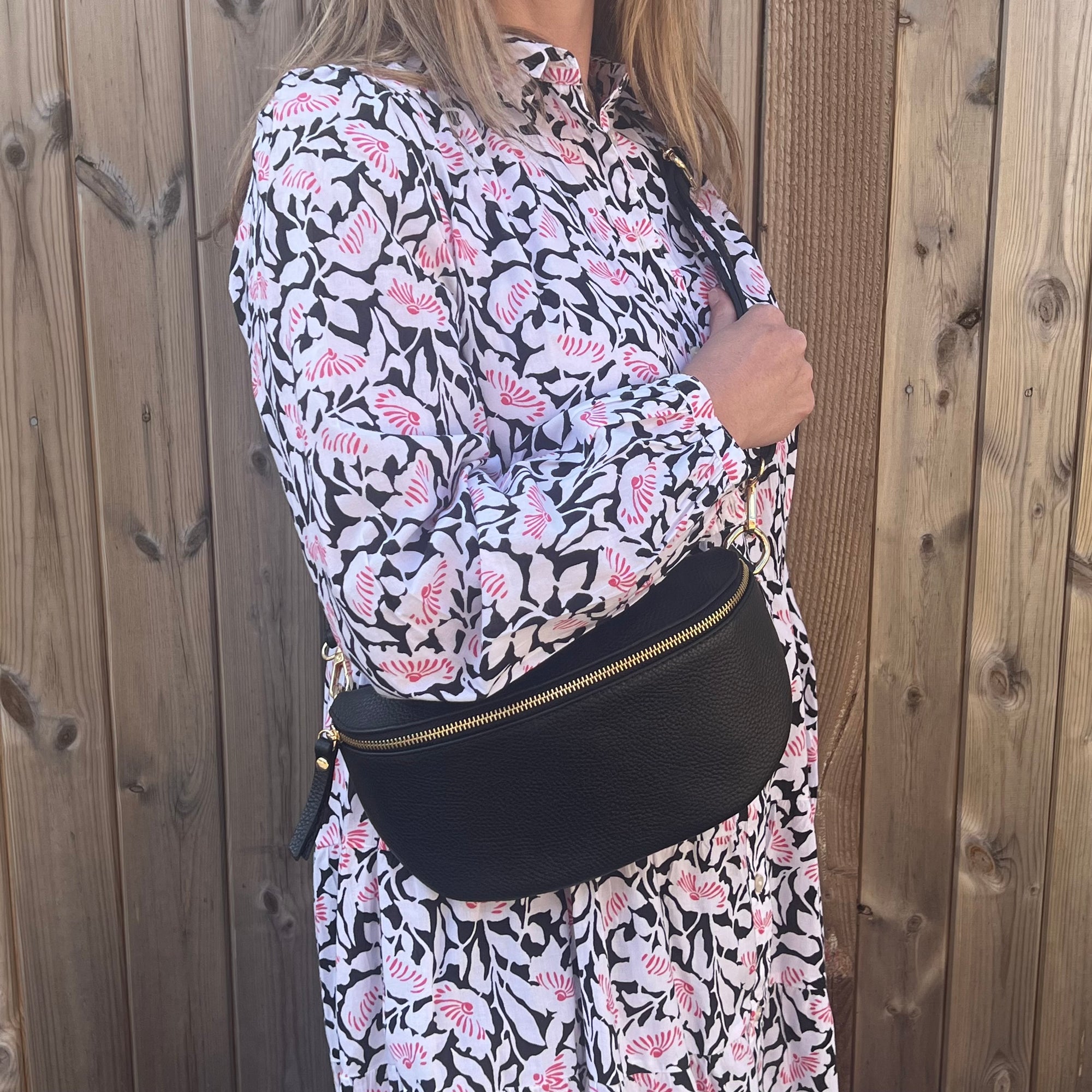 A woman wearing  a large black leather crossbody bag with an adjustable strap and gold hardware. 
