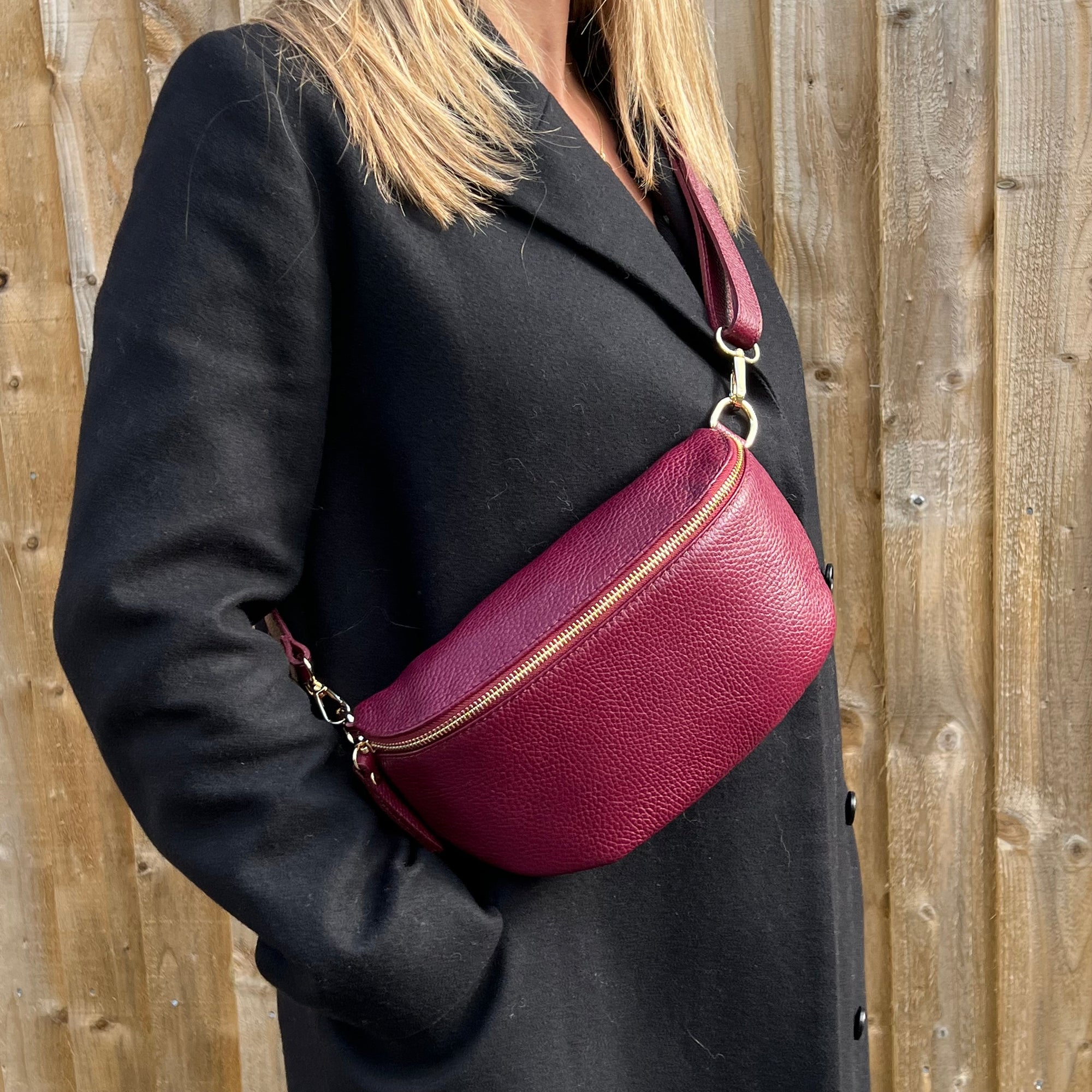 A person wearing a black coat is carrying a burgundy  leather crossbody bag with gold hardware.  

