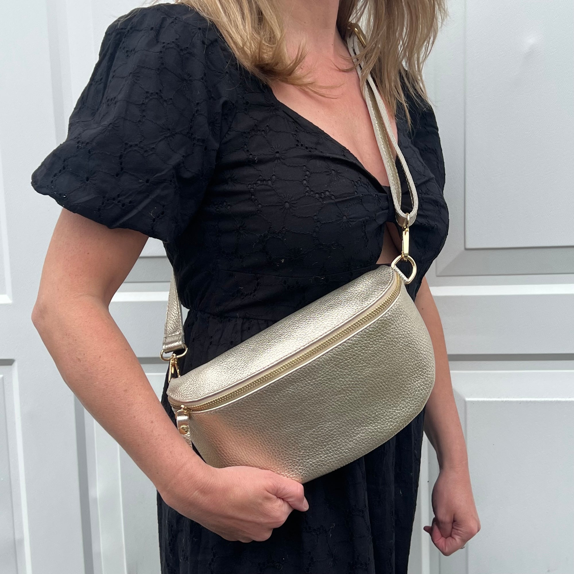 A woman wearing a black dress is holding a large gold leather crossbody bag with an adjustable strap.