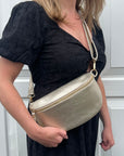 A woman wearing a black dress is holding a large gold leather crossbody bag with an adjustable strap.