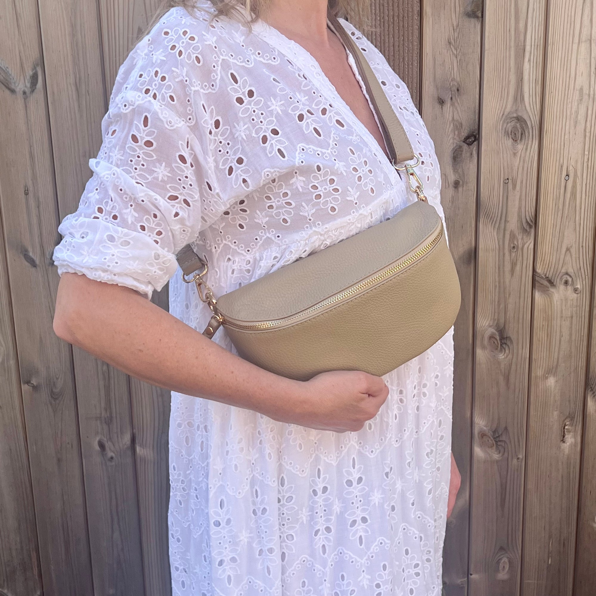 woman wearing a large light taupe crossbody/ waist bag  with and adjustable strap and gold hardware. 
